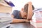 Tired disappointed schoolboy lowering his head sitting among pile of books, textbooks, school exercise books on his desk