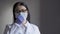 Tired doctor in mask on gray background. Brunette Asian woman corrects protective mask while looking at camera tiredly