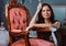 Tired female artist in apron holds paintbrush and leaning on a vintage chair in workshop.