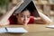 Tired frustrated teen asian girl makes roof with book on her head in library or room interior