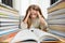 A tired girl with long blond hair holds her head next to a pile of textbooks and an open book.