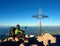 Tired hiker sit bellow crucifix on mountain peak. Iron cross at Alps mountain top.