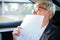 Tired mature businesswoman yawning while working on sales report documents, riding on a back seat of the car