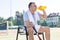 Tired mature man drinking from bottle while sitting on chair at tennis court on sunny day