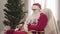 Tired Santa Clause sitting in rocking chair with decorated Christmas tree at the background. Exhausted young man in red