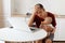 Tired sleepy handsome freelancer male wearing burgundy r shirt, posing in white kitchen, sitting in front of laptop with baby in