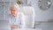 Tired sleepy old lady manager types on grey laptop at white table sitting on large chair against different shelves