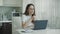 Tired woman using laptop computer at home. Serious woman drinking tea on kitchen