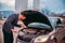 A tired worker is standing next to his car formally dressed and looking worried about the cars engine problem