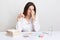 Tired young lady tries to concentrate on work, keeps hands on nose, holds spectacles, dressed in white formal shirt, poses at work