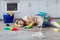 Tired young woman sleeping on floor at kitchen, surrounded by cleaning supplies, exhausted from housework