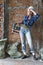 Tired young woman in work clothes and a protective helmet stands in front of a brick wall with a heavy hammer drill and wipes swea