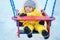 Toddler baby boy in a yellow snowsuit is swinging on a swing in a snow