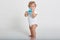 Toddler boy drinking water from blue feeding bottle, child wearing white bodysuit, looking away, standing barefoot against light