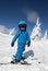 Toddler boy in helmet, goggles, blue overalls stands on skis on a snowy mountain slope