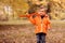 Toddler boy playing solo with orange toy airplane in autumn park. Trees with yellow and brown leaves in background