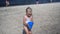 Toddler girl carrying toy bucket with water through beach sand. Blurred people