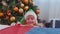 Toddler girl in Santa Claus Elf pajama hiding behind bed, playing hide and seek game, Christmas tree