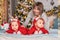 Toddler twins in red reindeer santa claus costumes are sitting at home with their mother against background of christmas tree