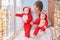 Toddler twins in red reindeer santa claus costumes are sitting at home with their mother against background of window with