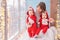 Toddler twins in red reindeer santa claus costumes are sitting at home with their mother against background of window with