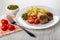 Tomato, fork on napkin,bowl with green peas, plate with cooked pasta, fried patties, green peas, tomato on wooden table