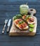 Tomato, mozzarella and basil sandwiches on dark wooden chopping board, pesto jar, dinnerware over black background