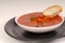 Tomato, red pepper, basil soup in white bowl with bread on a light gray background