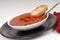 Tomato, red pepper, basil soup in white bowl with bread and spoon on a light gray background