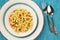 Top down, flat lay of delicious, vegetarian fusilli pasta with basil pesto, sweet corn, carrot and peas in a plate on a turquoise