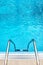 Top step and chrome hand rails of a swimming pool ladder and blue pool water background