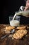 Top view of chocolate chip cookies, crumbs, spoon and hand with bottle pouring milk into glass cup, on wooden table and black back