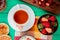 top view of cup of tea with white chocolate on teabag and bowl of strawberries with crispbreads and peach jam on green background