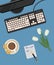 Top view of a desk background. There is a monitor, keyboard, a cup of coffee with cookies and white tulips on a blue background