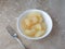 Top view of a fork next to a white bowl filled with canned pears atop a beige mottled tabletop