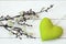 Top view of fuzzy willow branches and a small heart-shaped green plush on a white wooden background