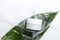 Top view of glass jar of facial cream on the fresh green natural leaf, shiny plastic wrapping material on the white surface.Empty