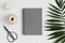 Top view of a grey notebook mockup with  workspace accessories and a palm leaf on a white table