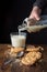 Top view of hand pouring milk with bottle on glass, chocolate chip cookies, crumbs, spoon on wooden table, black background,