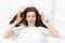 Top view of head of tired brunette young woman lying in bed with white sheet, pillow, blanket. Shocked female cover ears