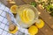 Top view of lemonade in a glass jug/pitcher with lemon slices. Lemons and wooden citrus pestle on table with textiles outdoors