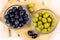 Top view of many green and black marinated whole olives in the glass bowls on light wooden board background in the kitchen.