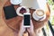 Top view mockup image of hands holding white smartphone with blank screen , tablet , laptop , coffee cup and cake on wooden table