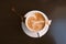 Top view of a mug of latte coffee with art on foam on dark wood background.