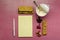 Top view of opened cup of yoghurt and spoon in it, heaps of nuts, raisins, diet bars, empty yellow notebook, pen on the pink surfa