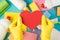 Top view photo of hands in yellow rubber gloves holding red paper heart multicolor sponges viscose rags garbage bags brush and