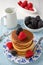 Top view of plate with pancakes, spoon, blackberries and raspberries, on blue striped cloth with white jug and bowls with blackber