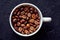 Top view of roasted whole coffee beans in white porcelain cup on gray textured background