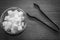 Top view of sugar cubes in a glass bowl with a pair of kitchen tongs foreceps kept on wooden floor. Contrasty black and white