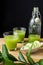 Top view of two glasses with cucumber juice and bottle, bowl with slices, mint and unfocused green branch, on wooden table and bla